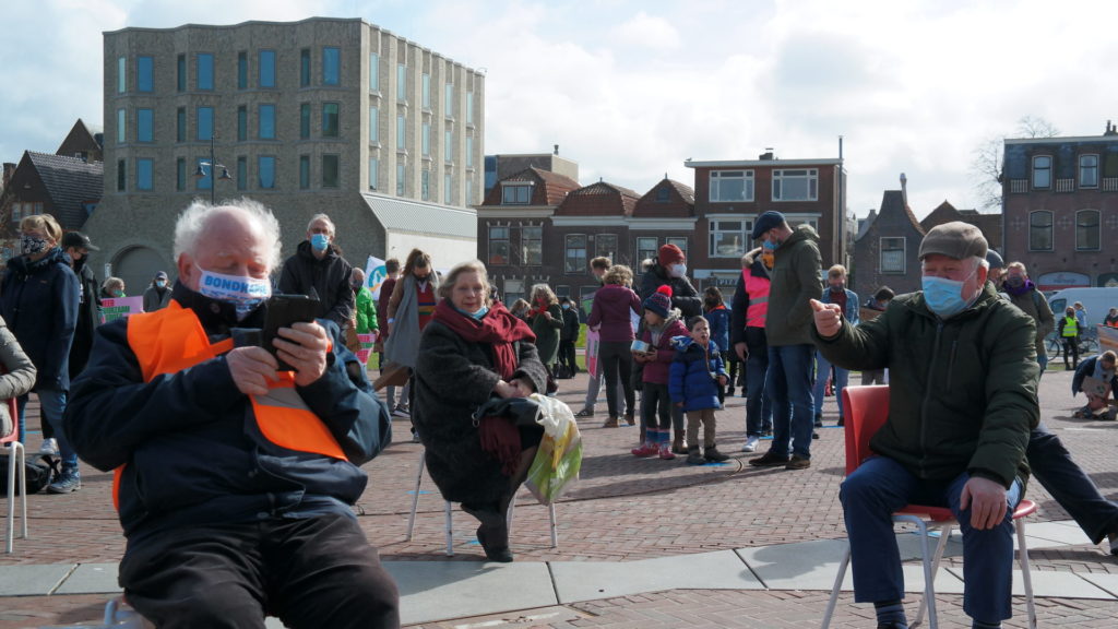 Op de voorgrond een drietal oudere, op stoeltjes zittende demonstranten op een plein. Erachter veel andere demonstranten, waaronder ook gezinnen met kinderen.