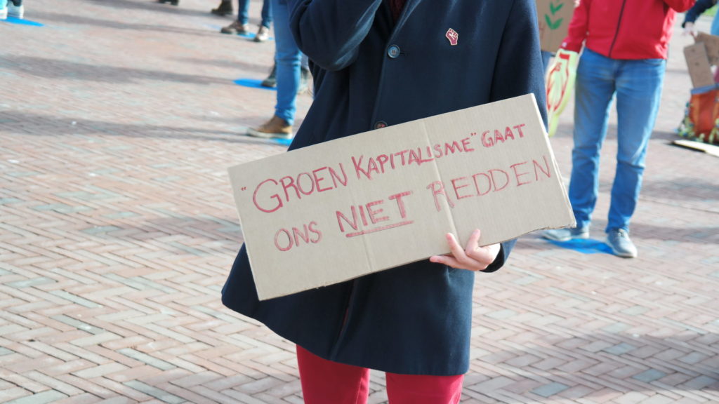 Je ziet enkel het middelste deel van een persoon en het bord wat diegene vasthoudt. De tekst luidt: "Groen kapitalisme gaat ons niet redden".