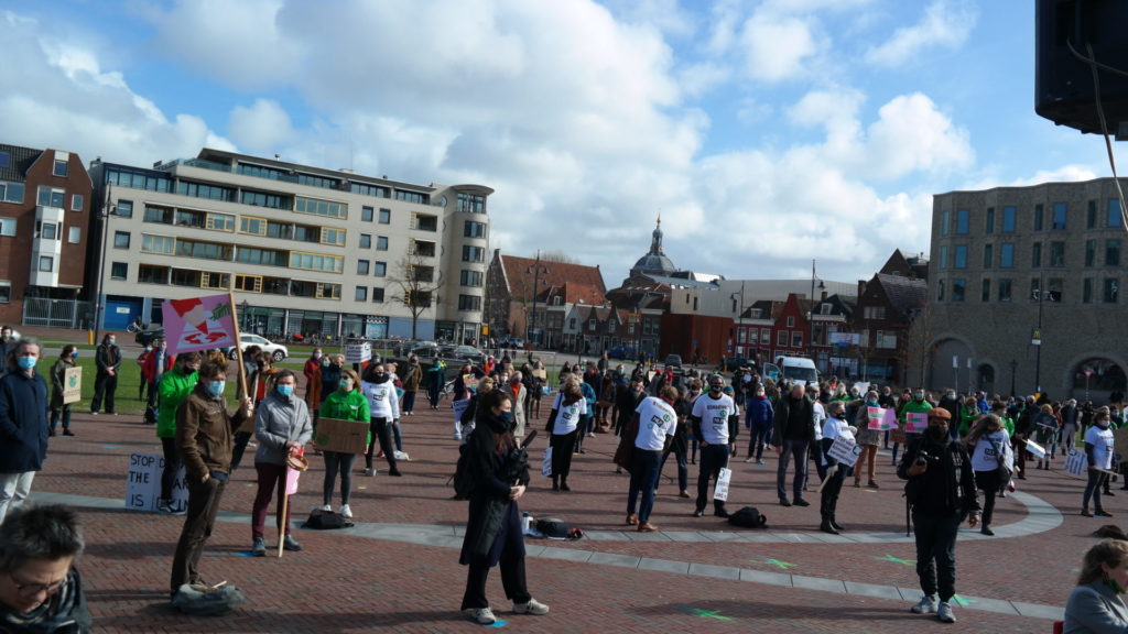 Een plein met tientallen demonstranten. Wegens corona houden ze afstand van elkaar. De zon schijnt, maar aan mensen hun kleding zie je dat het niet warm is.