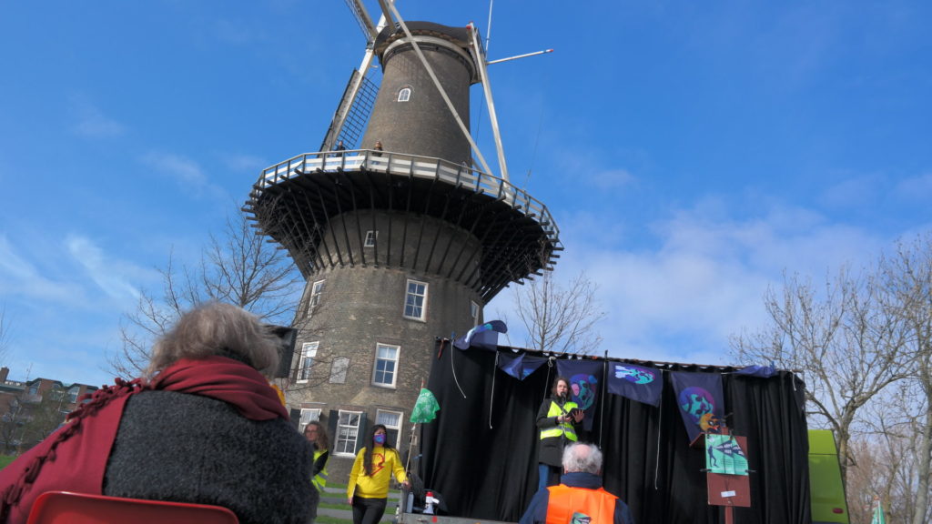 Bij een oud-Hollandse molen staat iemand achter een microfoon te spreken. De persoon heeft een veiligheidsjasje aan. Er zijn vlaggen te zien met een verdrietig kijkende aardbol.
