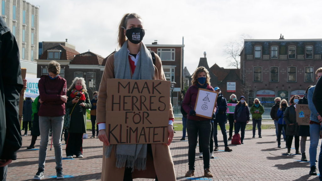 Een persoon met een lange lichtbruine jas en lange donkerblonde haren draagt een mondkapje waar "Black Lives Matter" op staat en een bord met de tekst: "Maak herres för klimatet".