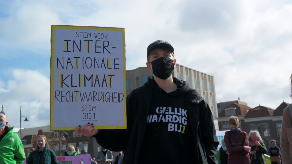 Een persoon met een petje op en een zwart mondkapje draagt een trui waar "Gelijkwaardig BIJ1" op staat. De persoon houdt een bord vast met de tekst "Stem voor internationale klimaatrechtvaardigheid - stem BIJ1".