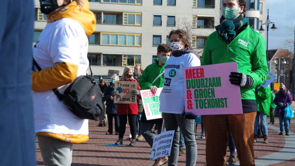 Een persoon met een jasje van Groen Links houdt een bord vast met daarop de tekst: "Meer duurzaam & groen & toekomst". Op de achtergrond een persoon met een bord met de tekst "There is no planet B".