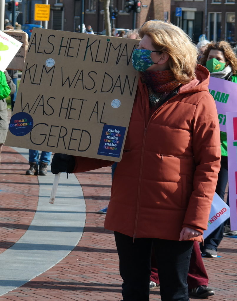 Een persoon met lange blonde haren, een vrolijk gekleurd mondkapje en een roestkleurige gewatteerde jas staat op een zonnige plek en houdt een bord vast met daarop de tekst "Als het klimaat KLM was, dan was het al gered".