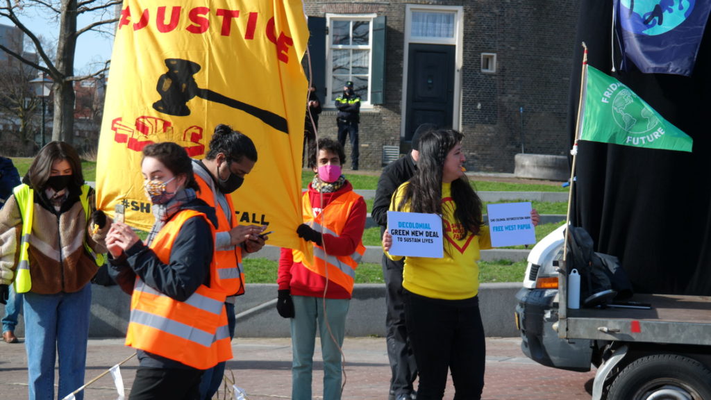 Enkele demonstranten staan bij het anti-Shell-spandoek. Eentje houdt een klein bord vast met de tekst "Decolonial Green New Deal to sustain lives".