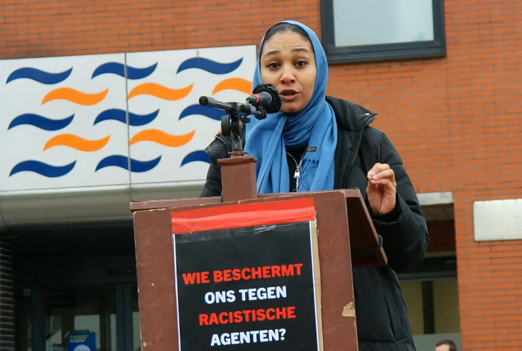 Saïda Ouarirou achter de microfoon. Ze draagt een blauwe hijab. Ze kijkt recht in de camera terwijl ze spreekt.