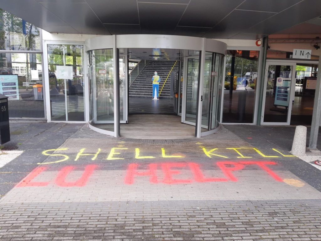 Voor de entree van een modern gebouw staat de tekst 'Shell kills - LU helpt'.