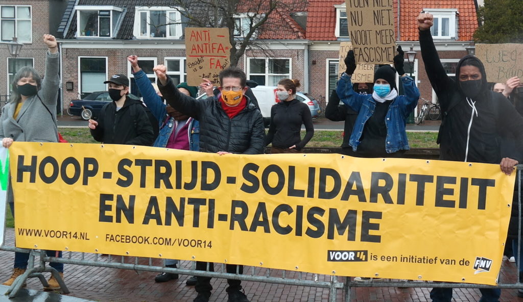 Demonstranten met een geel spandoek met de tekst "hoop - strijd - solidariteit en anti-racisme" en verwijzigingen naar Voor 14. Op de achtergrond meer demonstranten met verschillende borden, tegen een achtergrond van oude, typisch Nederlandse huisjes.