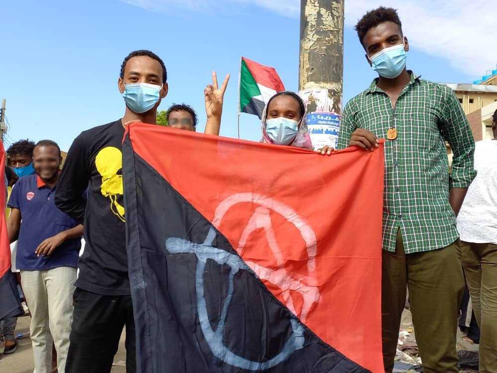 Een demonstratie. Drie demonstranten tonen een rood en zwarte vlag met een witte anarchy A. Op de achtergrond wappert de Soedanese vlag.