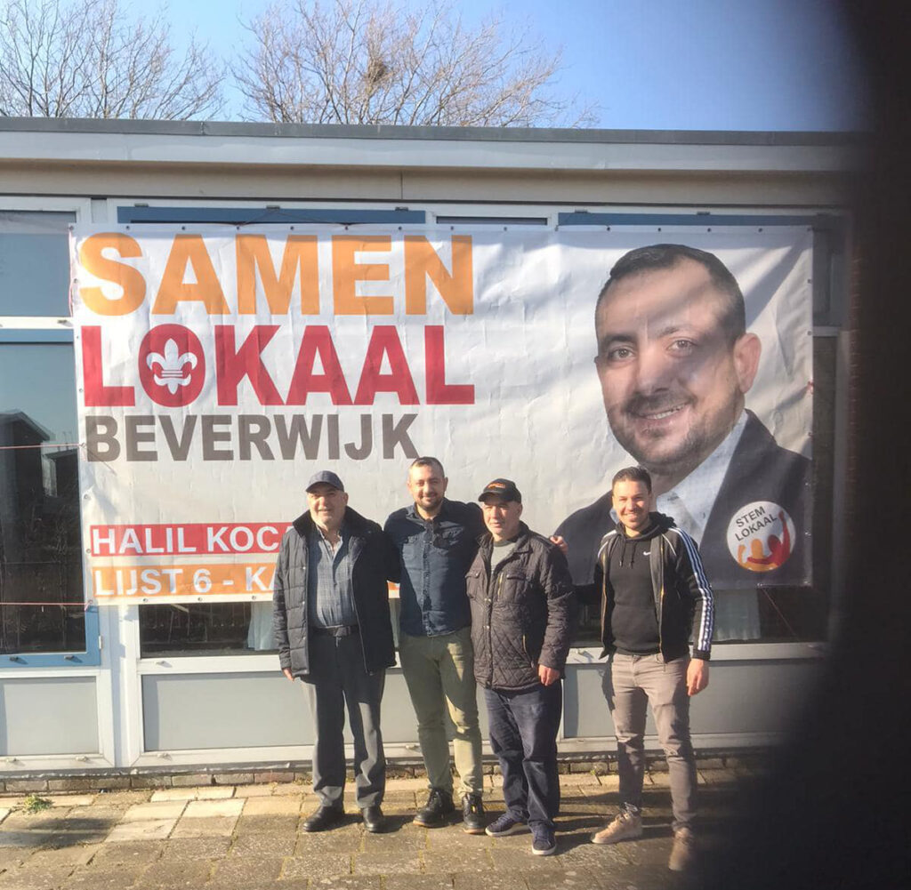 Vier mensen poseren voor het grijze wolven pand. Onder hen zijn Koçak en Bal. Aan het pand achter hen hangt een spandoek met de tekst: samen lokaal beverwijk, en een grote foto van Halil Koçak.