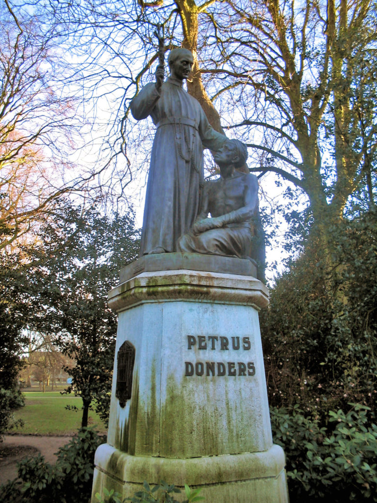 Het standbeeld van Petrus (Peerke) Donders. Je ziet een religieus gekleed persoon die een kruis omhoog houdt terwijl voor hem een Zwarte, halfnaakte man knielt en naar hem opkijkt.