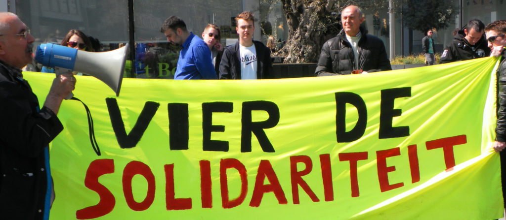 Een groot fluoriserend geel spandoek met de tekst "Vier de solidariteit". Op de voorgrond een persoon met een megafoon.