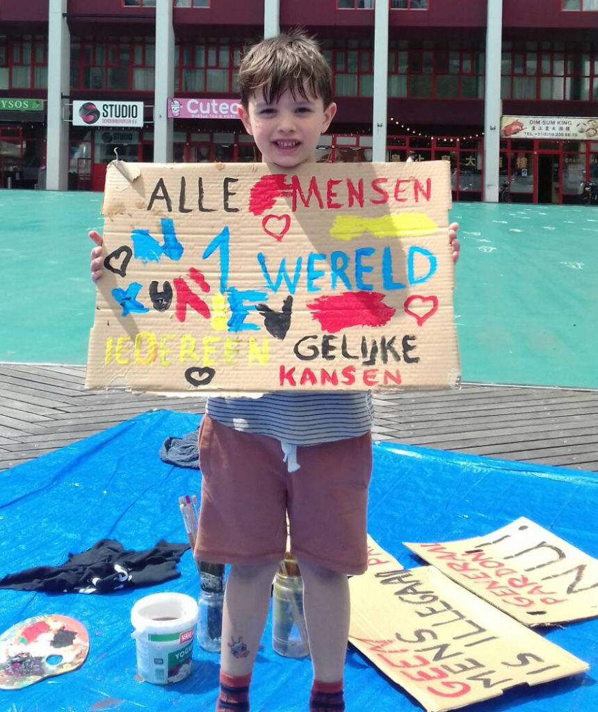 Een kind met een zelf beschilderd kartonnen bord. Er staat op: "Alle mensen, 1 wereld, iedereen gelijke kansen", eromheen allemaal hartjes.