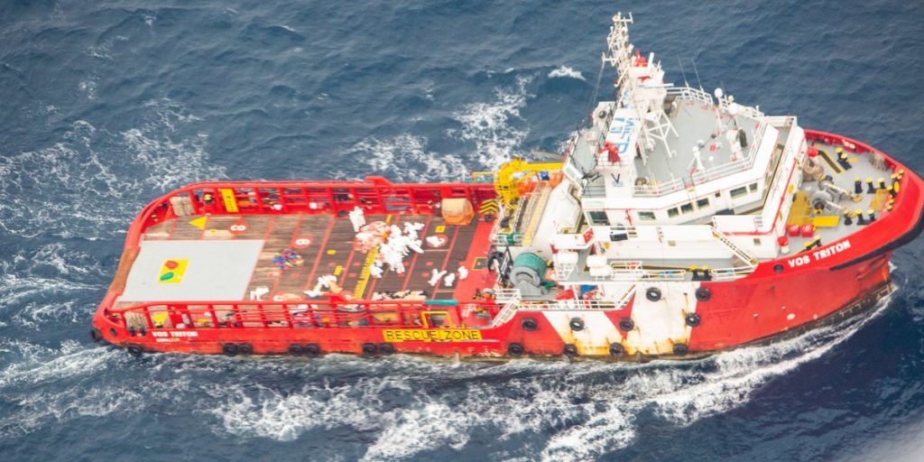 Foto van het rood met witte schip Vos Triton. Je ziet mensen in witte pakken op het dek.  Het gaat om een schip dat normaal gesproken boorplatformen verankert aan de zeebodem.