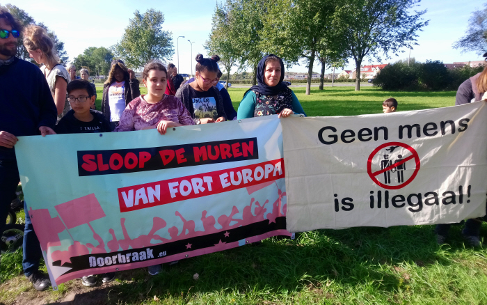 Demonstranten op een zonnig grasveld met spandoeken met de teksten "Geen mens is illegaal!" en "Sloop de muren van fort Europa".